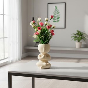 May include: A beige ceramic vase shaped like a person sitting in a cross-legged meditation pose with hands pressed together. The vase is filled with a bouquet of small artificial flowers in shades of pink, white, and green foliage.