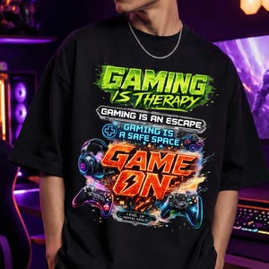 May include: Black graphic t-shirt with the text "GAMING IS THERAPY" and "GAME ON". The design includes images of headphones and game controllers. The shirt is worn by a person wearing sunglasses.