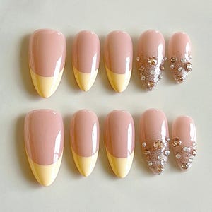 May include: A set of ten almond-shaped press-on nails featuring a glossy finish. Six nails have a two-tone design with a dusty rose base and a pale yellow tip. Four nails feature a dusty rose base with a glittery gold gradient and clear rhinestones.