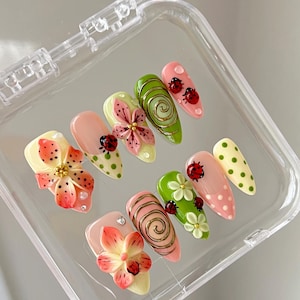 K&ouml;nnte beinhalten: Ein Set aus zehn k&uuml;nstlichen Fingern&auml;geln mit verschiedenen Designs, darunter 3D-Blumen in Rosa und Orange, gr&uuml;ne Spiralen mit goldenen Akzenten, rote Marienk&auml;fer sowie hellgr&uuml;ne oder rosafarbene Punkte, in einem transparenten Kunststoffetui.