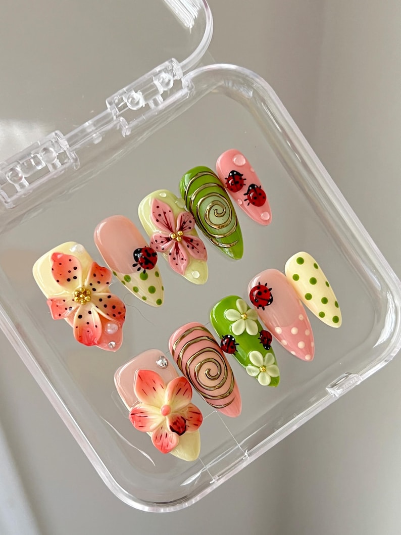 K&ouml;nnte beinhalten: Ein Set aus zehn k&uuml;nstlichen Fingern&auml;geln mit verschiedenen Designs, darunter 3D-Blumen in Rosa und Orange, gr&uuml;ne Spiralen mit goldenen Akzenten, rote Marienk&auml;fer sowie hellgr&uuml;ne oder rosafarbene Punkte, in einem transparenten Kunststoffetui.