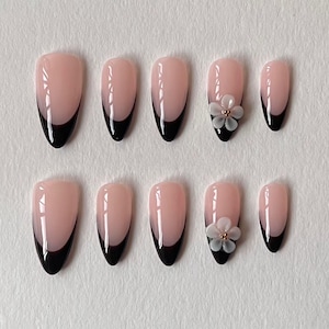 May include: A set of ten almond-shaped press-on nails featuring a glossy nude base with black French tips. Two of the nails are accented with a small, white, five-petaled flower embellishment centred with tiny gold-toned beads at the nail tip.