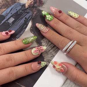 K&ouml;nnte beinhalten: Lange, mandelf&ouml;rmige Fingern&auml;gel mit verschiedenen 3D-Designs, darunter rosa und gr&uuml;ne Blumenmuster, goldene Metallspiralen und kleine rote Marienk&auml;fer-Applikationen. Die Manik&uuml;re verwendet eine Palette aus zartem Rosa, Hellgr&uuml;n und Wei&szlig;.