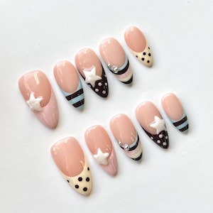 Puede incluir: Un juego de diez u&ntilde;as postizas en forma de almendra con base color nude y varios dise&ntilde;os, incluyendo rayas horizontales negras y azul claro, puntos negros sobre puntas crema y estrellas blancas en relieve con peque&ntilde;os adornos met&aacute;licos plateados.