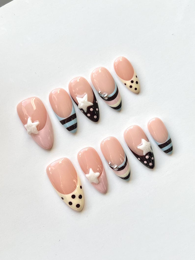 Puede incluir: Un juego de diez u&ntilde;as postizas en forma de almendra con base color nude y varios dise&ntilde;os, incluyendo rayas horizontales negras y azul claro, puntos negros sobre puntas crema y estrellas blancas en relieve con peque&ntilde;os adornos met&aacute;licos plateados.