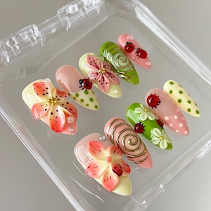 K&ouml;nnte beinhalten: Ein Set aus zehn k&uuml;nstlichen Fingern&auml;geln in einer transparenten Kunststoffbox mit verschiedenen Designs, darunter rosa und orangefarbene 3D-Blumen, gr&uuml;ne Wirbel, rote Marienk&auml;fer-Akzente und Punkte in Rosa, Gr&uuml;n und Creme mit goldenen Details.