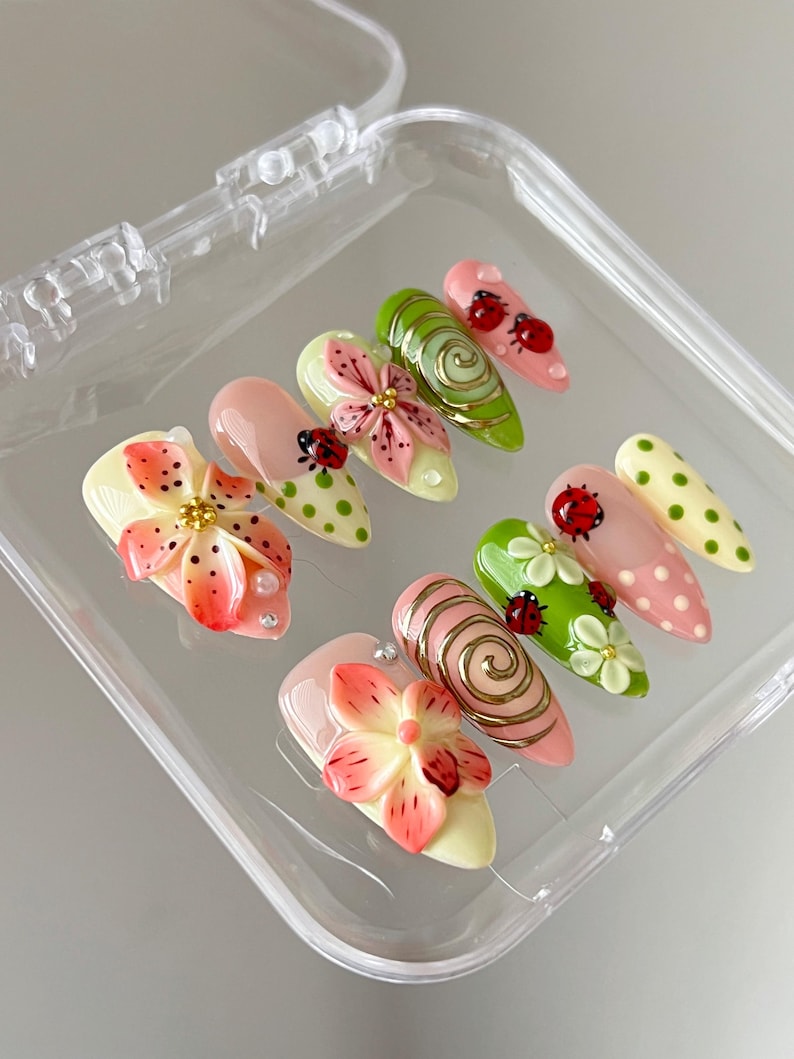 K&ouml;nnte beinhalten: Ein Set aus zehn k&uuml;nstlichen Fingern&auml;geln in einer transparenten Kunststoffbox mit verschiedenen Designs, darunter rosa und orangefarbene 3D-Blumen, gr&uuml;ne Wirbel, rote Marienk&auml;fer-Akzente und Punkte in Rosa, Gr&uuml;n und Creme mit goldenen Details.