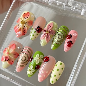 Op de afbeelding: Een set van tien amandelvormige kunstnagels met diverse ontwerpen, waaronder 3D-bloemen in roze en oranje, rode lieveheersbeestjes, groene en roze stippen en gouden wervelpatronen, gepresenteerd in een doorzichtig plastic opbergdoosje.