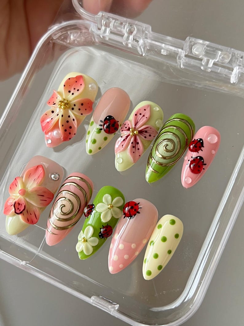 K&ouml;nnte beinhalten: Ein Set aus zehn mandelf&ouml;rmigen Press-on-N&auml;geln mit verschiedenen Designs, darunter 3D-Blumen in Pink und Orange, rote Marienk&auml;fer, gr&uuml;ne und rosafarbene Punkte sowie goldene Wirbelmuster, pr&auml;sentiert in einem transparenten Kunststoffetui.