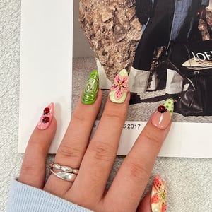 K&ouml;nnte beinhalten: Mandelformige Kunstn&auml;gel mit verschiedenen Designs, darunter eine gr&uuml;ne Spirale, eine rosa Blume, Marienk&auml;fer auf rosa und hautfarbenen Untergr&uuml;nden sowie ein Blumenmuster. Die N&auml;gel werden mit einem silberfarbenen Doppelring getragen.