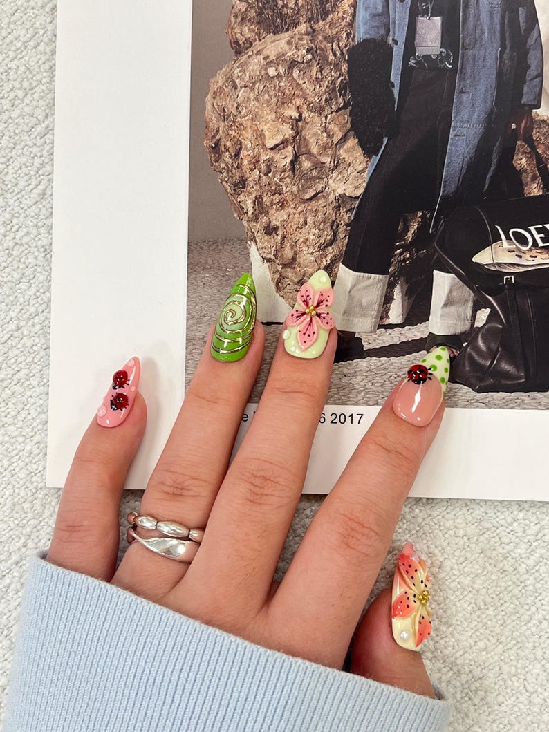 K&ouml;nnte beinhalten: Mandelformige Kunstn&auml;gel mit verschiedenen Designs, darunter eine gr&uuml;ne Spirale, eine rosa Blume, Marienk&auml;fer auf rosa und hautfarbenen Untergr&uuml;nden sowie ein Blumenmuster. Die N&auml;gel werden mit einem silberfarbenen Doppelring getragen.