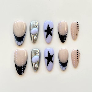 Könnte beinhalten: Ein Set aus zehn mandelförmigen Press-on-Nägeln mit verschiedenen Designs. Einige Nägel haben eine nudefarbene Basis mit schwarz-weißen Punktmustern, andere sind metallic-silber mit helllila Cabochons und einige zeigen schwarze Sterne auf Lavendel.