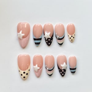 Puede incluir: Un juego de diez u&ntilde;as postizas en forma de almendra con base color nude, decoradas con lunares negros, rayas horizontales en negro y azul claro, dijes en forma de estrella blanca y peque&ntilde;as esferas met&aacute;licas plateadas en algunas u&ntilde;as.