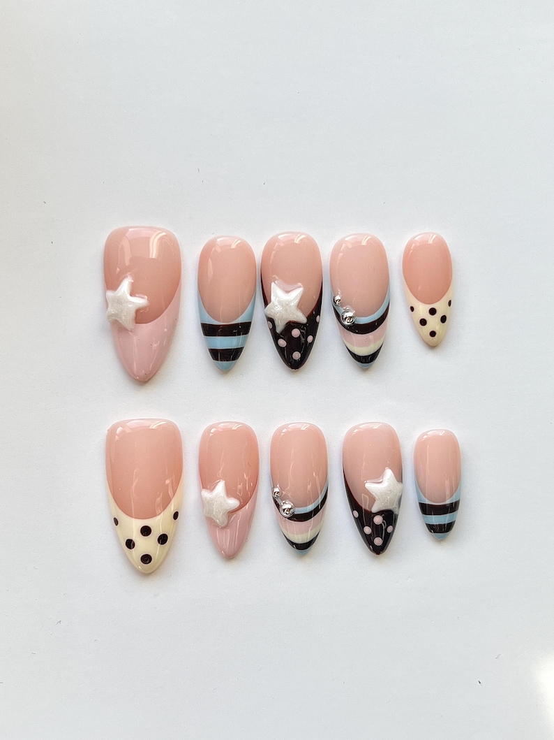 Puede incluir: Un juego de diez u&ntilde;as postizas en forma de almendra con base color nude, decoradas con lunares negros, rayas horizontales en negro y azul claro, dijes en forma de estrella blanca y peque&ntilde;as esferas met&aacute;licas plateadas en algunas u&ntilde;as.