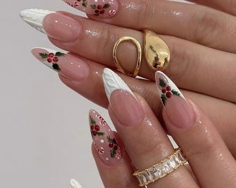 May include: Almond-shaped nails featuring a nude base with white French tips, some with a raised cable-knit texture. The design includes hand-painted red holly berries with green leaves and small candy canes. Two gold rings are worn on the fingers.