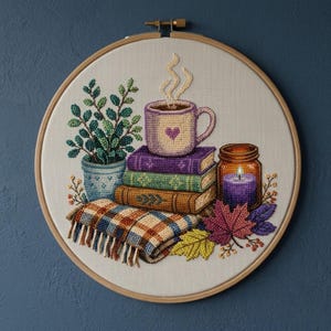 May include: A diamond painting kit in a circular wooden embroidery hoop featuring a mug with a heart on a stack of three books, a potted plant, a plaid blanket, a lit purple candle in a glass jar, and colorful autumn leaves on white fabric.