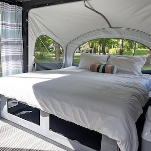 May include: A pop-up camper interior with a bed covered in a light gray striped comforter and pillows. The camper has large arched windows offering a view of the outdoors. A striped curtain hangs on the side.