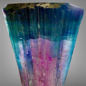 May include: A raw, vertically striated tourmaline crystal featuring a distinct color gradient. The top section displays deep blue and teal hues, transitioning into a vibrant magenta pink at the base, with a flat, dark, metallic-looking top surface.