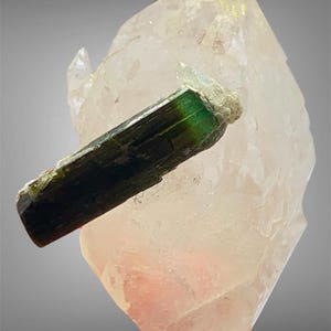 May include: A raw, elongated green tourmaline crystal with a dark, opaque base transitioning to a translucent emerald hue at the tip, resting horizontally against a large, pale, semi-translucent quartz cluster with a jagged, crystalline structure.