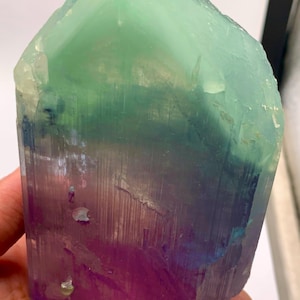 May include: A large, translucent, multi-colored crystal specimen featuring a distinct color gradient. The top section is a pale mint green, transitioning into a deep blue middle, and ending in a vibrant purple base. The crystal has a vertical, striated texture.
