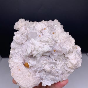 May include: A cluster of white crystalline mineral formations featuring a rough, textured surface with small, embedded orange-colored inclusions scattered throughout the structure, creating a jagged and irregular natural geological specimen.