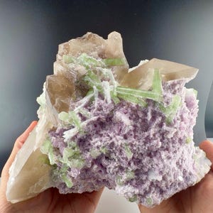 May include: A mineral specimen featuring a cluster of translucent, light brown quartz crystals alongside vibrant, pale green prismatic crystals, all set against a textured, light purple lepidolite matrix with small white crystalline inclusions.