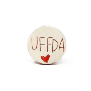 May include: White ceramic round ornament with the word "UFFDA" written in brown lettering and a red heart.
