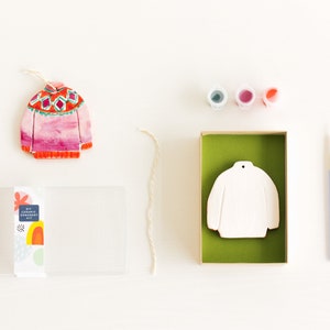 May include: A DIY ceramic ornament kit featuring a white ceramic sweater ornament, three small bottles of paint, a paintbrush, and a length of twine. The kit also includes a pre-painted sweater ornament with a pink, red, and green pattern.