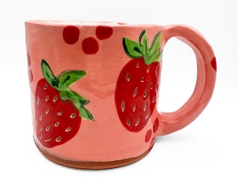 May include: A ceramic mug with a pink exterior, featuring hand-painted red strawberries and red polka dots. The mug has a rounded handle and a brown base. The strawberry design includes green leaves and small white details.