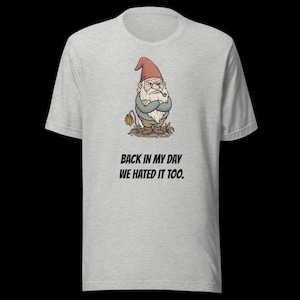 May include: Light grey t-shirt with a grumpy gnome illustration wearing a red hat and holding a pipe. The text "BACK IN MY DAY WE HATED IT TOO" is printed below the gnome. The shirt is made of a soft material.