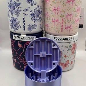 Puede incluir: Varios tarros de comida aislados de acero inoxidable, cada uno con una capacidad de 740 ml. Los tarros presentan diseños florales y de lazos en rosa, azul y blanco. Un tarro está abierto, revelando una tapa morada. El texto "FOOD JAR 25oz" es visible.