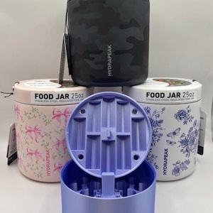 Puede incluir: Tres tarros térmicos de acero inoxidable Hydrapeak de 25oz y un recipiente de plástico morado claro con tapa abatible. Los tarros presentan un patrón de camuflaje negro, lazos rosas y un diseño floral azul con el texto FOOD JAR 25oz.