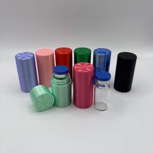 May include: A collection of cylindrical storage containers in various colours including purple, pink, red, green, blue, and black, alongside a clear glass vial with a blue cap, featuring textured starburst patterns on the flat top lids.