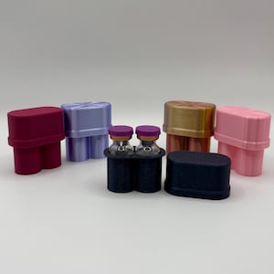 Op de afbeelding: Een verzameling van zes ovaalvormige houders met twee sleuven voor kleine glazen flesjes. De houders zijn uitgevoerd in diverse effen kleuren, waaronder donkerroze, lichtpaars, metallic goud, lichtroze en donkerblauw, met twee flesjes erin geplaatst.