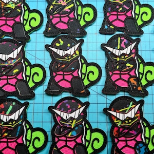 May include: A collection of embroidered patches featuring a cartoon turtle character wearing white sunglasses. The turtle has a black body with pink and neon green accents, including a bright green shell and colorful paint splatter details throughout.