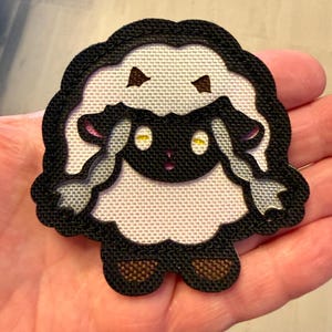 May include: A fabric patch featuring an embroidered cartoon sheep with a black face, yellow eyes, and white wool. The sheep has long, flowing white hair, brown hooves, and is outlined with a thick black border. The design is flat and textured.