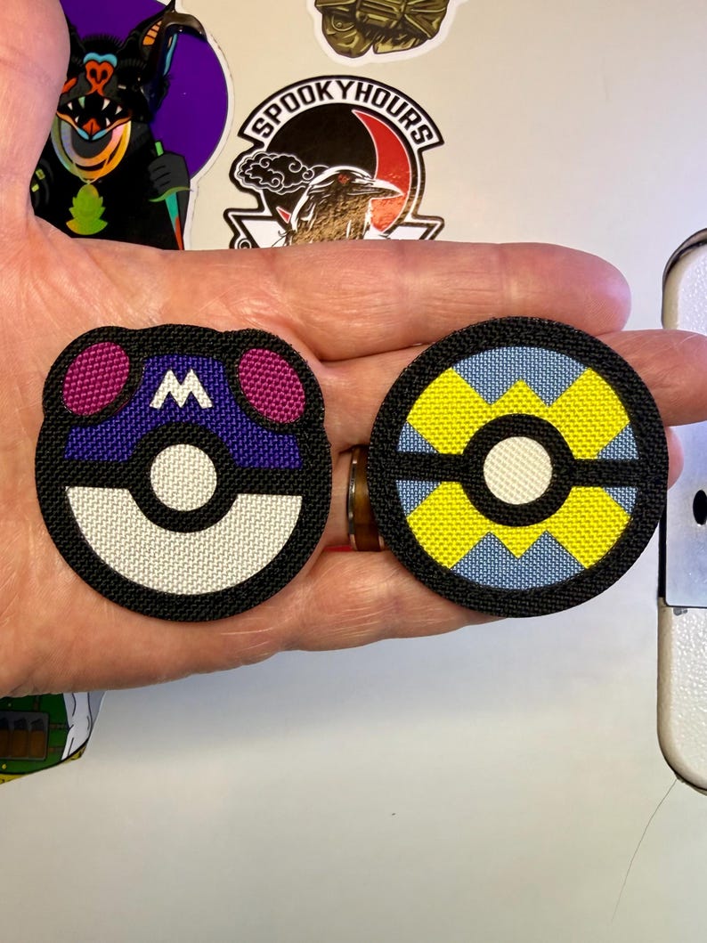May include: Two circular embroidered patches featuring stylised designs. The left patch is purple, white, and black with a white letter M. The right patch is yellow, light blue, and black. Both patches have a central white circle and black borders.