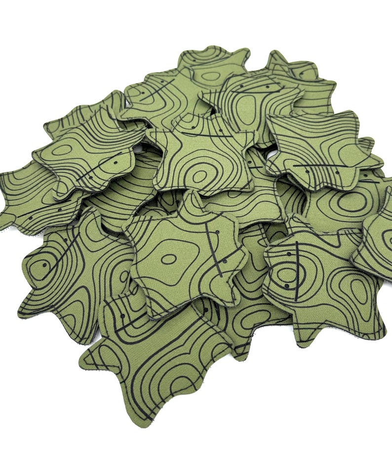 May include: A pile of green, star-shaped fabric items with black contour lines. The items have a unique, organic shape and a topographic map design. The fabric has a slightly textured appearance, and the black lines create a detailed pattern.