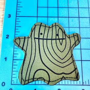 May include: A green and black fabric patch in the shape of a Ditto Pok&eacute;mon. The patch has a wood grain pattern and a simple face with two black dots for eyes and a black line for a mouth. The patch is on a blue grid.