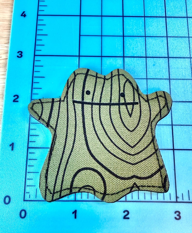 May include: A green and black fabric patch in the shape of a Ditto Pok&eacute;mon. The patch has a wood grain pattern and a simple face with two black dots for eyes and a black line for a mouth. The patch is on a blue grid.