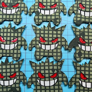 May include: A collection of nine embroidered patches shaped like a cartoon creature with a wide grin, pointed ears, and spiky hair. Each patch features a dark olive green and black grid pattern, bright red eyes, and a white mouth with teeth.