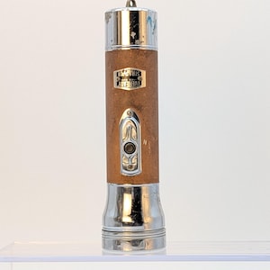 May include: A vintage Eveready Strapper flashlight with a brown textured body and polished chrome metal ends. The device features a central chrome switch plate and a small metal hanging loop at the top. The Eveready Strapper logo is visible.
