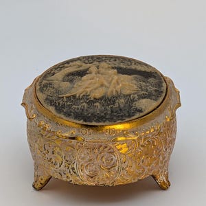 May include: A round, footed trinket box featuring a gold-toned exterior with intricate embossed floral patterns. The lid displays a circular, cream-colored relief carving of figures in a landscape set against a dark, textured background.