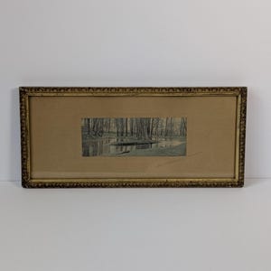 May include: A rectangular landscape print featuring a forest scene with a small stream, centered on a tan mat board within a decorative gold-toned frame. A handwritten signature is visible in the bottom right corner of the matting.
