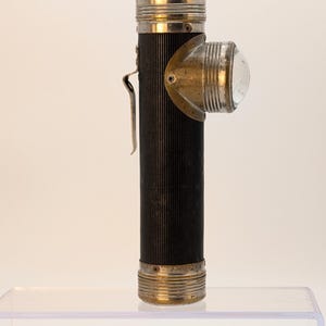 May include: A vintage right-angle flashlight with a black ribbed cylindrical body and metallic silver-toned ends. It features a prominent side-mounted circular glass lens housing with a brass-colored reflector and a metal belt clip on the side.
