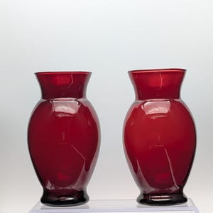 May include: Two matching ruby red glass vases with a bulbous body, a narrow neck, and a flared rim. The vases feature a smooth, glossy finish and are positioned side by side, showcasing a deep, translucent red color throughout the glass structure.