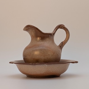 May include: A ceramic pitcher and matching bowl set featuring a speckled brown and tan glaze. The pitcher has a curved handle and a pouring spout, while the bowl has a scalloped edge design. Both pieces exhibit a consistent, textured finish.