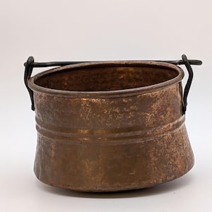 May include: A round, hammered copper pot featuring a weathered, dark brown patina. The vessel has a wide, open top and a single, thin, dark metal handle attached to two side loops, allowing the handle to pivot. The exterior surface is textured.
