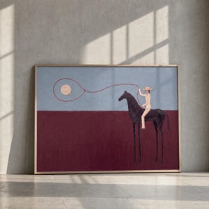 May include: A rectangular framed painting featuring a person in a wide-brimmed hat riding a black horse against a two-tone background. The upper section is light blue, while the lower section is deep maroon. A red lasso line loops in the sky.