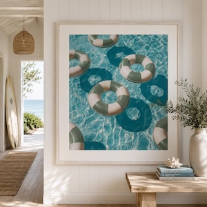 May include: A framed wall art print featuring a top-down view of multiple green and white striped pool floats drifting in clear blue water. The image captures the rippling water surface and the shadows cast by the circular floats below.